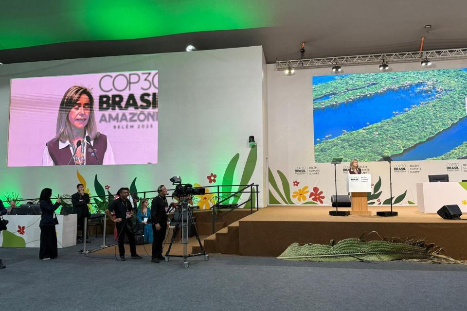 A woman speaks at a podium on stage during the COP30 Brazil Amazonia event, with cameras and attendees present and a large screen displaying her image.