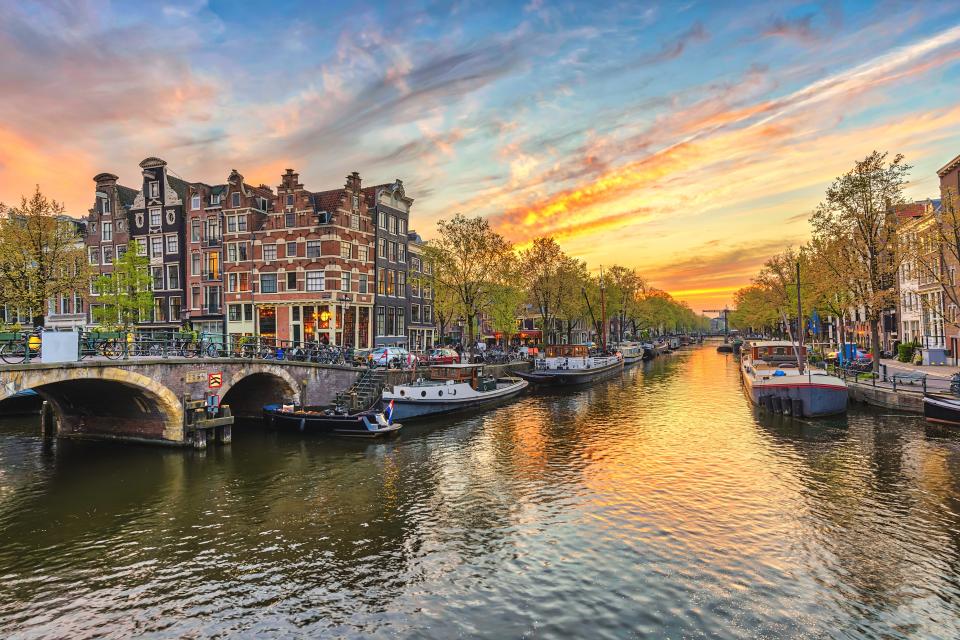 A canal in Amsterdam at sunset, with boats moored along the water, historic buildings, a stone bridge, and trees lining both sides of the canal.
