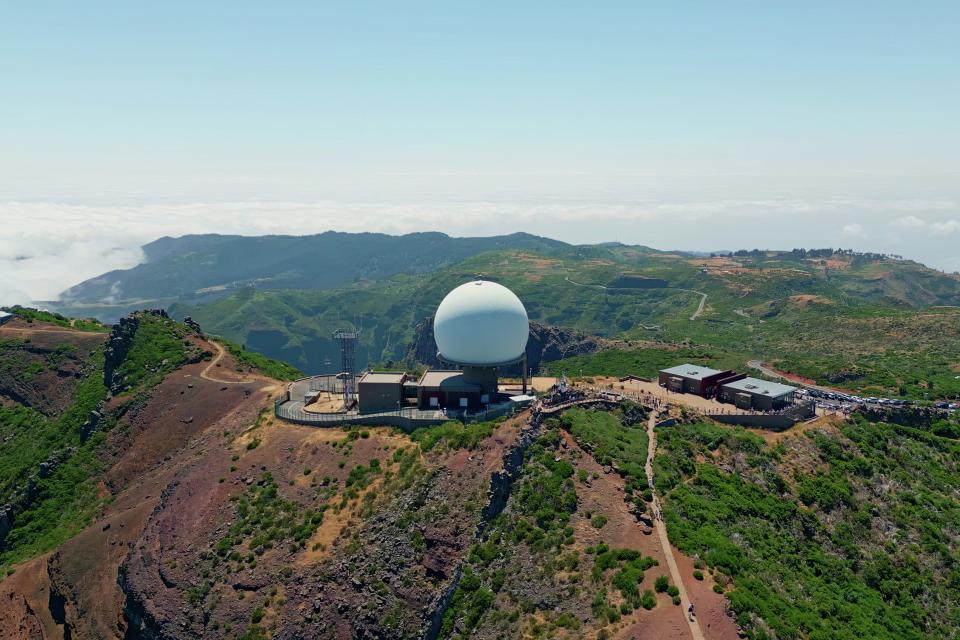 Large white radar dome facility situated on a mountain ridge surrounded by green hills, with scattered buildings and clear skies above.