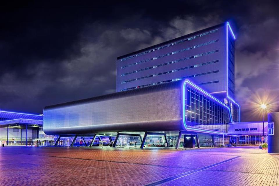 A modern building with blue neon lighting features a large, elevated structure supported by pillars, photographed at night with a cloudy sky in the background.